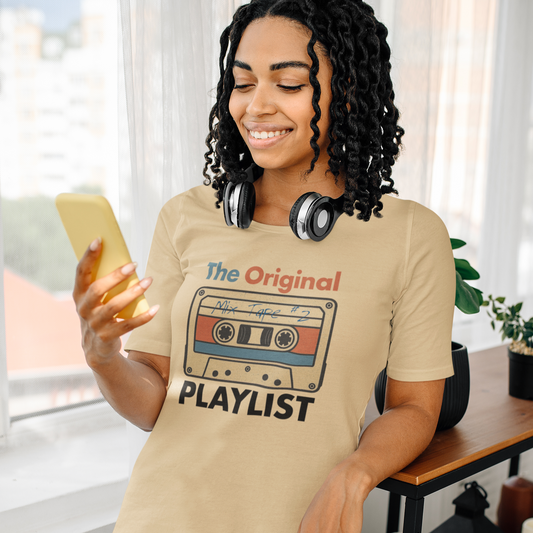 Woman wearing a beige t-shirt with a cassette tape graphic and text that says THE ORIGINAL PLAYLIST, holding a phone in a room with a window and plant.