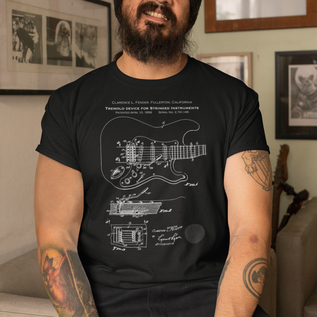 Man wearing a black t-shirt with a guitar design, sitting in a room with framed pictures on the wall.