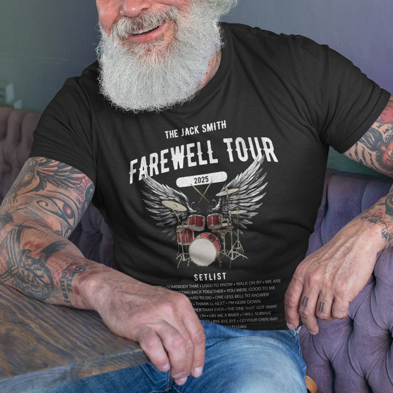 Man with tattoos wearing a black t-shirt with 'Farewell Tour' design, sitting on a purple couch.