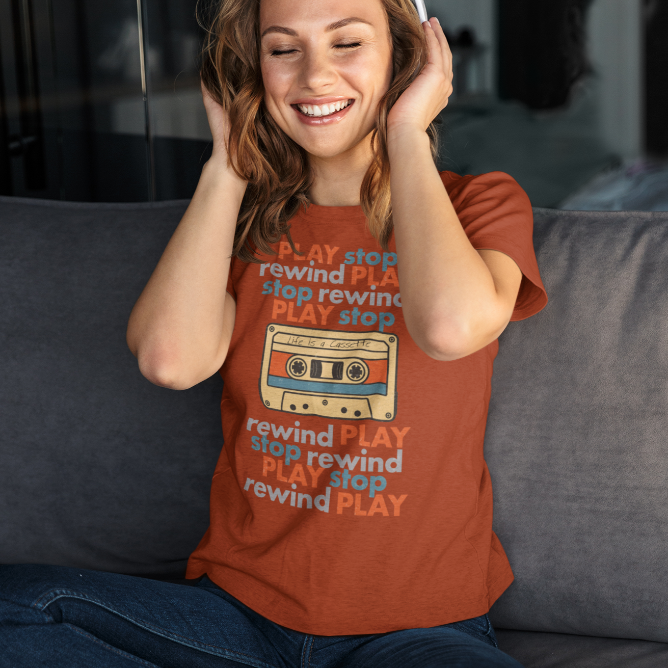 Woman wearing a red t-shirt with PLAY< STOP< REWIND text, and a graphic of a cassette tape with "Life is a Cassette" handwritten on it, sitting on a couch and listening to music.
