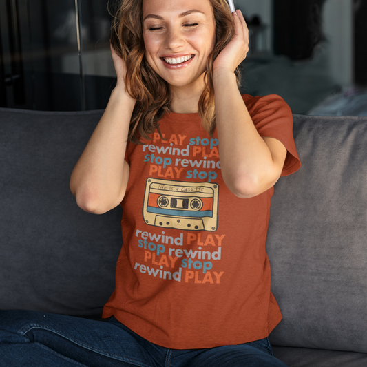 Woman wearing a red t-shirt with PLAY< STOP< REWIND text, and a graphic of a cassette tape with "Life is a Cassette" handwritten on it, sitting on a couch and listening to music.