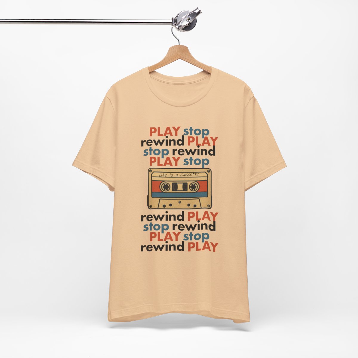 Beige t-shirt with cassette tape graphic and 'Play stop rewind' text on a hanger against a white background